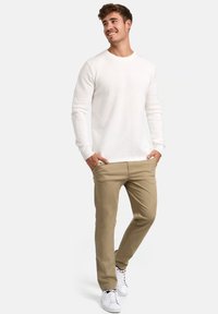 Weißer strukturierter Strickpullover mit langen Ärmeln, kombiniert mit beigen Chinos und weißen Sneakers. Das Modell steht mit einer Hand in der Tasche und lächelt.