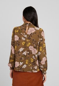 Brun blus med blommigt mönster, lång ärm, rynkad krage och nyckelhålsöppning i ryggen. Har rosa och gula blommor på ett strukturerat tyg.