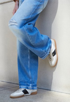 Persoon draagt lichtblauwe gescheurde jeans en witte sneakers met een zwarte streep, staand met één been gebogen tegen een beige muur.