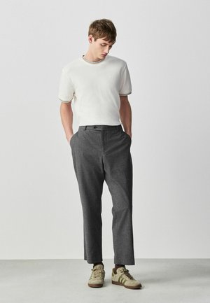 Junger Mann steht und blickt nach unten, trägt ein weißes Kurzarmhemd mit Besatz, graue Hose und beige Sneakers, Hände in den Taschen.