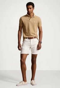 Man i beige polo, vita slitna shorts, brun flätad bälte och beige sneakers som står mot en enfärgad bakgrund.