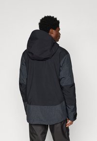 Bărbat îmbrăcat într-o jachetă cu glugă de culoare neagră și gri închis, cu un model în grilă pe mâneci și pe partea inferioară a spatelui, stând de profil și privindu-se spre dreapta.