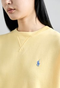Sudadera amarilla con cuello redondo y detalles de costura visibles. Presenta un pequeño logo azul en la parte frontal. Textura suave, diseño informal.