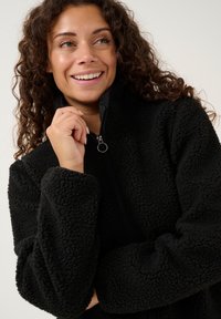 Svart fleecejacka med dragkedja i kragen, strukturerad yta och breda manschetter, med en minimalistisk design och mjukt, fluffigt material.