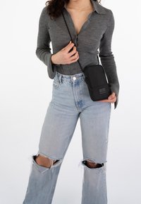 Svart axelremsväska med rektangulär form, slät yta och dragkedjeslutning. Bärs med en grå långärmad tröja och slitna ljusa jeans.