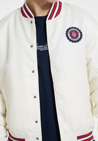 Weißes Bomberjackett mit rot-blauen gestreiften Rippkragen und -bündchen. Verfügt über Druckknöpfe und ein kreisförmiges gesticktes Logo auf der Brust.