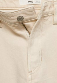 I pantaloni in denim beige presentano un bottone e una cerniera argentati, con cuciture chiare e una superficie liscia. La vita ha un dettaglio minimale.