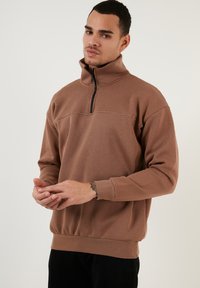 Sudadera de cuello con cremallera en marrón, fabricada en un tejido suave, que presenta un dobladillo y puños acanalados. Diseño limpio con una textura sutil. Sin patrones ni acentos visibles.