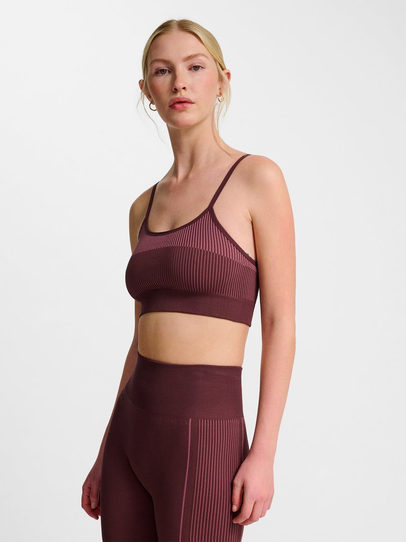 Ung kvinde med blondt hår iført matchende ribbet mørkerød sports-bh og højtaljede leggings mod ensfarvet lyst baggrund.