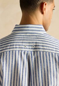 Sarkā ar svītrām, kas sastāv no zilām un baltām svītrām, ar pogu lejas daļu, krokotu muguru ar diviem metāla caurumiem un teksturētu audumu.