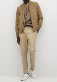 Tan suedebomberjakke over en beige striktrøje, parret med beige tapered bukser og hvide sneakers. Simpelt design og neutrale farver.