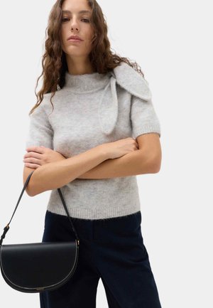 Donna con capelli lunghi e ricci indossa un maglione grigio a maniche corte con dettaglio a fiocco e pantaloni scuri, tenendo una borsa nera con cuciture bianche.
