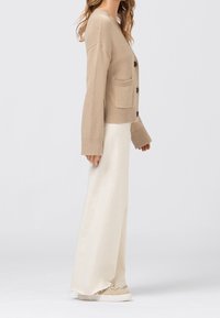 Kvinna i beige knappad cardigan, lång vit kjol och beige sneakers, stående mot en enfärgad ljus bakgrund i profil.