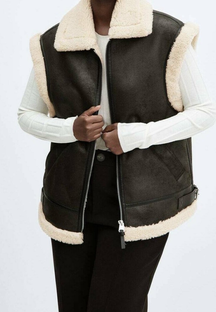 Personne portant une veste sans manches en peau retournée marron foncé avec col et bordure en fourrure crème, par-dessus un haut blanc à manches longues, fermant la fermeture éclair à l'avant.