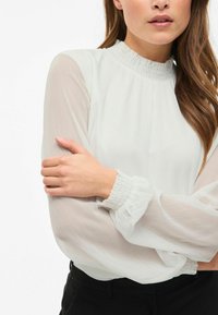 Hvid bluse med en høj, samlet halsudskæring og gennemsigtige ærmer. Elastiske manchetter og blødt, tekstureret stof. Parret med sorte bukser.