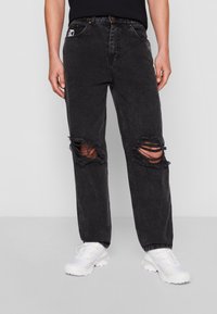 Jeans negros desgastados con grandes detalles rasgados en las rodillas, hechos de mezclilla; combinados con zapatillas deportivas blancas. Cuenta con un parche de logo en el lateral.