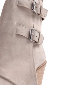 Μποτάκι αστραγάλου από μπεζ σουέντ με δύο ασημένιες αγκράφες και πελώριο ξύλινο τακούνι, σε κοντινή λήψη λεπτομέρειας.