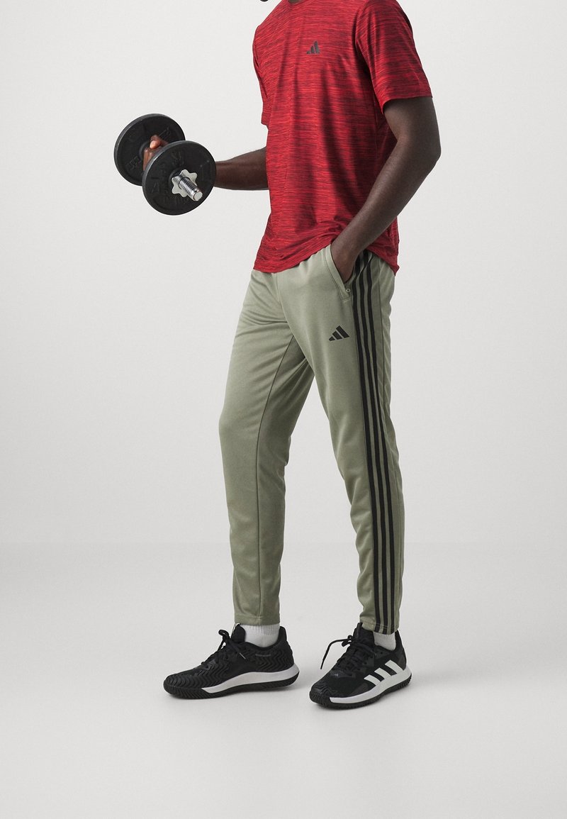Man in een rood Adidas shirt, olijfgroene Adidas trainingsbroek en zwarte Adidas sneakers, met een dumbbell in één hand, staande tegen een witte achtergrond.