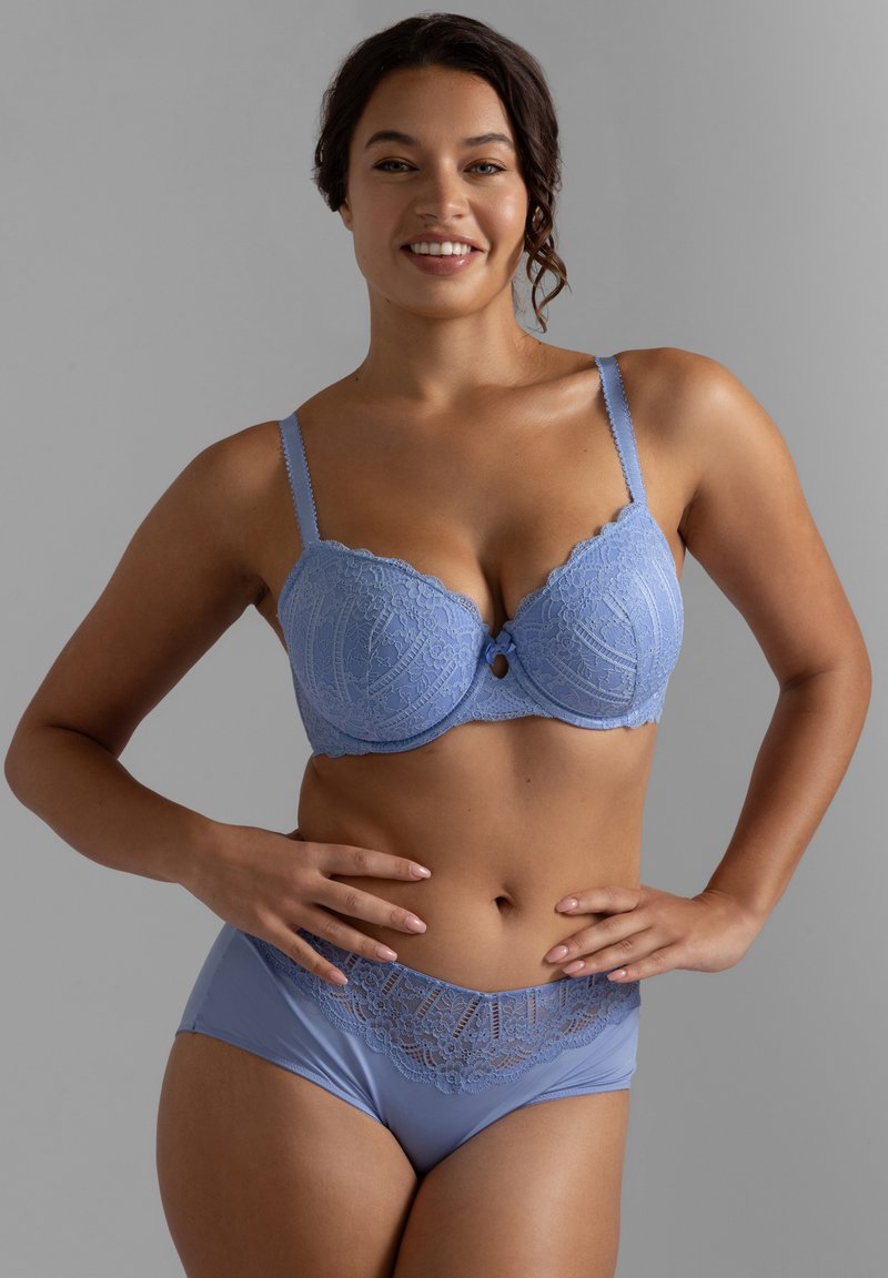 Χαμογελαστή γυναίκα που φοράει ταιριαστό σουτιέν από δαντέλα σε ανοιχτό μπλε χρώμα και εσώρουχα, στέκεται με τα χέρια στη μέση μπροστά σε γκρι φόντο.