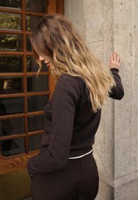 Brūns adīts džemperis ar garām piedurknēm, ar baltu apmali ap apakšmalu. Fona attēlā redzams teksturēts pelēks sienas un koka durvis.