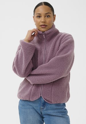 Femme aux cheveux tirés en arrière, portant des boucles d'oreilles en perles et une veste en polaire lavande zippée, posant avec une main près du visage et les bras croisés.