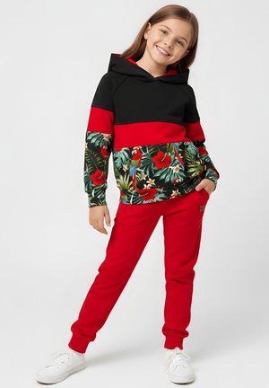 Uśmiechnięta dziewczyna stoi z jedną ręką w kieszeni, ubrana w czarno-czerwoną bluzę z kapturem z tropikalnym kwiatowym i papuzim nadrukiem, czerwone spodnie i białe trampki.