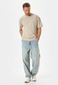 Beige tekstureret t-shirt med korte ærmer, parret med lyseblå baggy jeans og hvide sneakers. Simpelt, afslappet design.