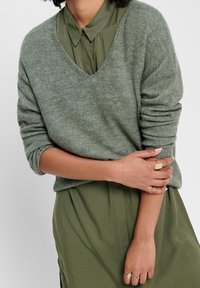 Henkilö, joka käyttää vihreää kauluspukua löysän harmaavihreän V-painanteen neuleen alla, ja heidän sormissaan on kerroksittain kultasormuksia.