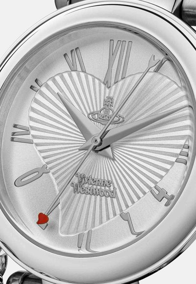 Montre analogique argentée avec un cadran texturé en forme de cœur, des chiffres romains et un accent cœur rouge sur l'aiguille des secondes. Nom de la marque affiché.