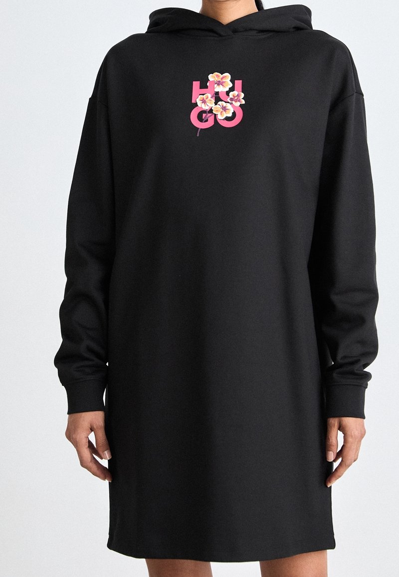 Personne portant une robe à capuche noire avec le texte rose « HUGO » et de petites fleurs roses sur la poitrine, sur un fond uni clair.