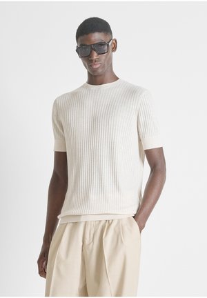 Hombre con suéter de manga corta crema acanalado, pantalones beige plisados y grandes gafas de sol negras, de pie con una mano en el bolsillo.