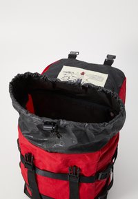 Sac à dos rouge avec un dessus noir, fermeture à cordon de serrage et matériel réglable. L'intérieur présente une étiquette graphique. Matériau texturé avec des sangles.