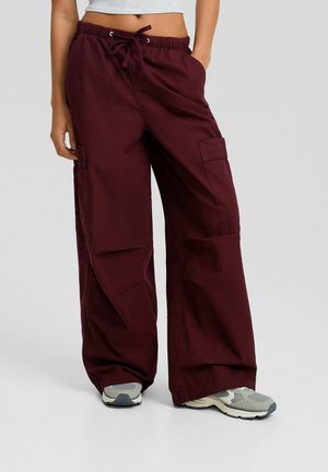 Osoba nosząca burgundowe spodnie cargo o szerokich nogawkach z sznurkiem w talii i szare sneakersy, stojąca z jedną ręką w kieszeni.