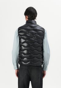 Gilet imbottito nero con un motivo trapuntato, caratterizzato da un colletto alto e una texture lucida, indossato sopra una camicia azzurro chiaro e jeans scuri.