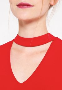 Abito rosso con dettagli a collana, caratterizzato da un scollo a V. Tessuto liscio e leggero con un design pulito e minimalista. Vista ravvicinata della zona del collo.