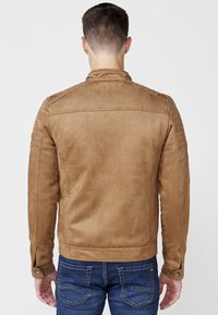 Hnedá umelá semišová bunda s prešívanými detailami na pleciach, s gombíkovými manžetami a slim fit strihom, skombinovaná s modrými džínsami.