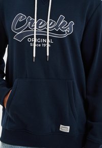Sudadera azul marino de tela suave, con un gráfico blanco que dice "Creeks ORIGINAL", cordones ajustables y bolsillo frontal con etiqueta.