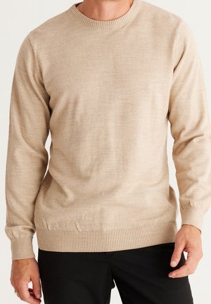 Homme portant un pull en maille beige à manches longues et un pantalon noir, debout devant un fond uni.