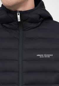Fekete puffdzseki cipzáros elől, kapucnival, vízszintes varrással és az "ARMANI EXCHANGE" logóval a mellkason.
