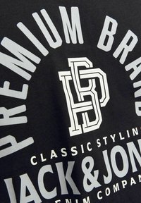 Juodas medvilnės marškinėliai su dideliu baltu tekstu, įskaitant "PREMIUM BRAND" ir "JACK & JONES". Grafikos logotipas centre. Lygus paviršius.