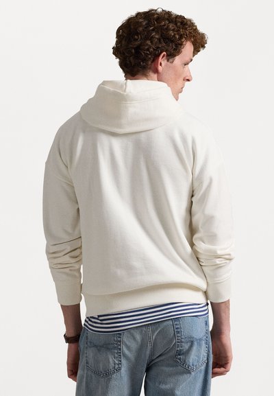 Κρέμα χρώματος φούτερ με μεγάλη, μαλακή κουκούλα, ριμπ μανσέτες και στρίφωμα. Φοράει πάνω από ένα ριγέ μπλε και λευκό πουκάμισο, συνδυασμένο με ανοιχτόχρωμα τζιν.