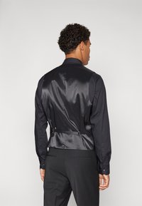 Schwarze Satin-Weste mit glatter Textur, verstellbaren Riemen auf der Rückseite und taillierter Passform, kombiniert mit einem klassischen schwarzen Hemd.