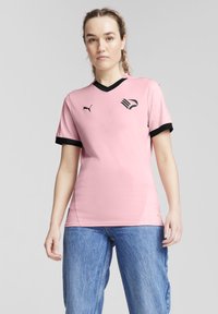 Ružový športový dres s čiernym véčkom a čiernymi manžetami na rukávoch. Logo Puma na ľavej hrudi s grafiku vtáka. Hladká textúra látky.
