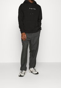 Hombre vestido con una sudadera negra de Tommy Jeans, pantalones gris oscuro y zapatillas blancas, de pie contra un fondo claro y liso.