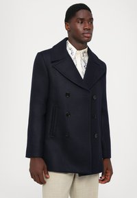 Námornícky kabát so zapínaním na dvojité gombíky, veľkými klopami, čiernymi gombíkmi a postrannými vreckami, vyrobený z textúrovanej tkaniny.