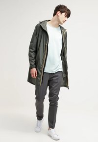 Khaki pláštěnka se žlutým zipem, světle modré tričko, šedé kalhoty a bílé tenisky. Hladká látka, volný střih, otevřený design.