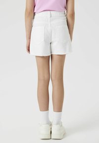 Shorts en denim blanc avec un ourlet effiloché, poches arrière et taille mi-haute. Portés avec des baskets blanches et des chaussettes côtelées, vus de dos.