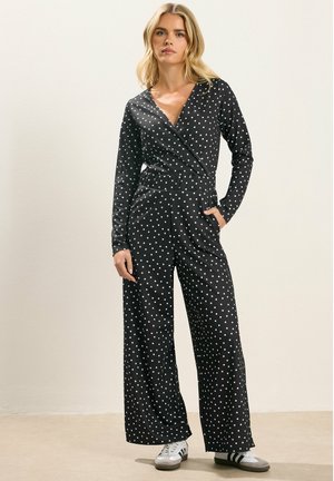 Kvinde står iført en sort langærmet polkaprikket jumpsuit med vide ben og hvide sneakers, den ene hånd i lommen.