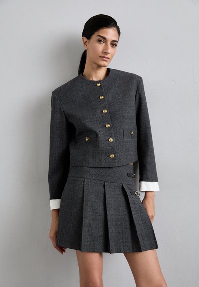 Grijze tweed cropped jas met gouden knopen, gecombineerd met een plissérok met accenthardware en contrasterende witte manchetten.