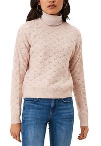 Pull en maille rose clair avec un motif en losanges, poignets et ourlet côtelés. Porté par-dessus un col roulé pâle et assorti à un jean bleu.
